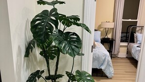 Una habitación con una planta de monstera en maceta, una cama con colcha blanca, una mesita de noche con lámpara y una ventana con cortinas.