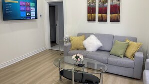 Una sala de estar moderna con un sofá gris, una mesa de centro de vidrio y un televisor de pantalla plana montado en la pared que muestra la interfaz de un servicio de streaming.