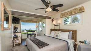 Ein Schlafzimmer mit Bett, Deckenventilator, Fenster mit Vorhängen, Nachttisch und Blick ins Freie.