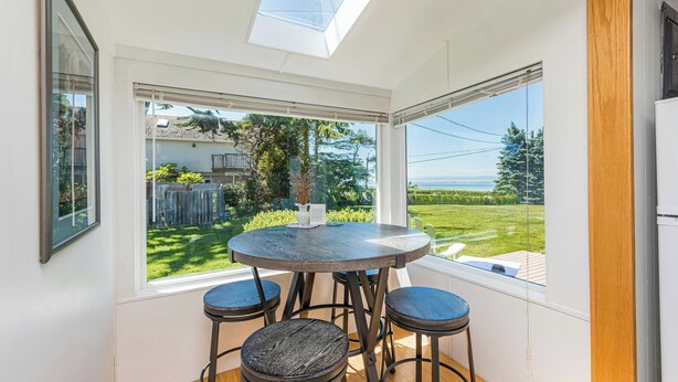 A room with a round table and four stools, large windows with a view of a backyard and a body of water, and a skylight above the table. - Lavender Suite - King, full kitchen, jetted tub (Sequim)