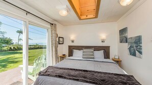 A bedroom with a large bed, wooden ceiling, two bedside tables, a nightstand, a view of a grassy area through a sliding door, and two wall-mounted lights. - Lavender Suite - King, full kitchen, jetted tub (Sequim)