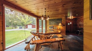 Un interior de madera con una mesa de comedor de cristal rodeada de bancos, una araña y vistas de un edificio rojo al exterior.