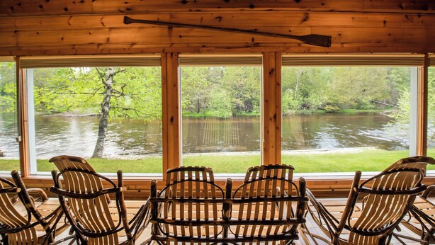 Un interior de madera con ventanas amplias que dan a un río, sillas de madera dispuestas al frente y techo de madera.