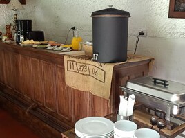 Disposición de buffet con cafetera, dispensador de zumo y un gran contenedor negro, probablemente para almacenamiento de alimentos.