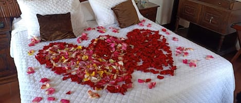 A bed with a quilted white bedspread and pillows, adorned with rose petals arranged in a heart shape.
