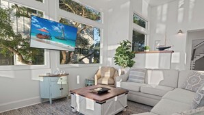 A modern living room with a large TV displaying a tropical beach scene, a beige sofa, a white armchair, a small side table, and a coffee table. - Marsh front home 10 min to beach and downtown- pet friendly! (Charleston)