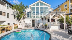 A swimming pool with a deck, a covered outdoor seating area, and a building with large windows in the background. - Marsh front home 10 min to beach and downtown- pet friendly! (Charleston)