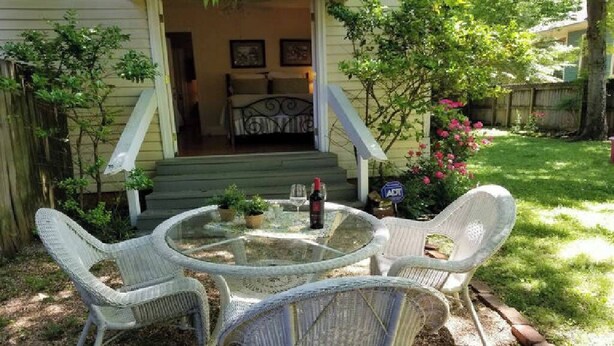Une terrasse avec des chaises en osier et une table en verre, un petit jardin entouré d’une clôture en bois et une entrée avec un escalier.