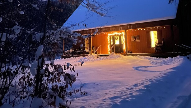 Schneebedeckte Landschaft mit einem Gebäude, das eine Veranda und einen Sitzbereich im Freien besitzt, beleuchtet von warmem Licht.