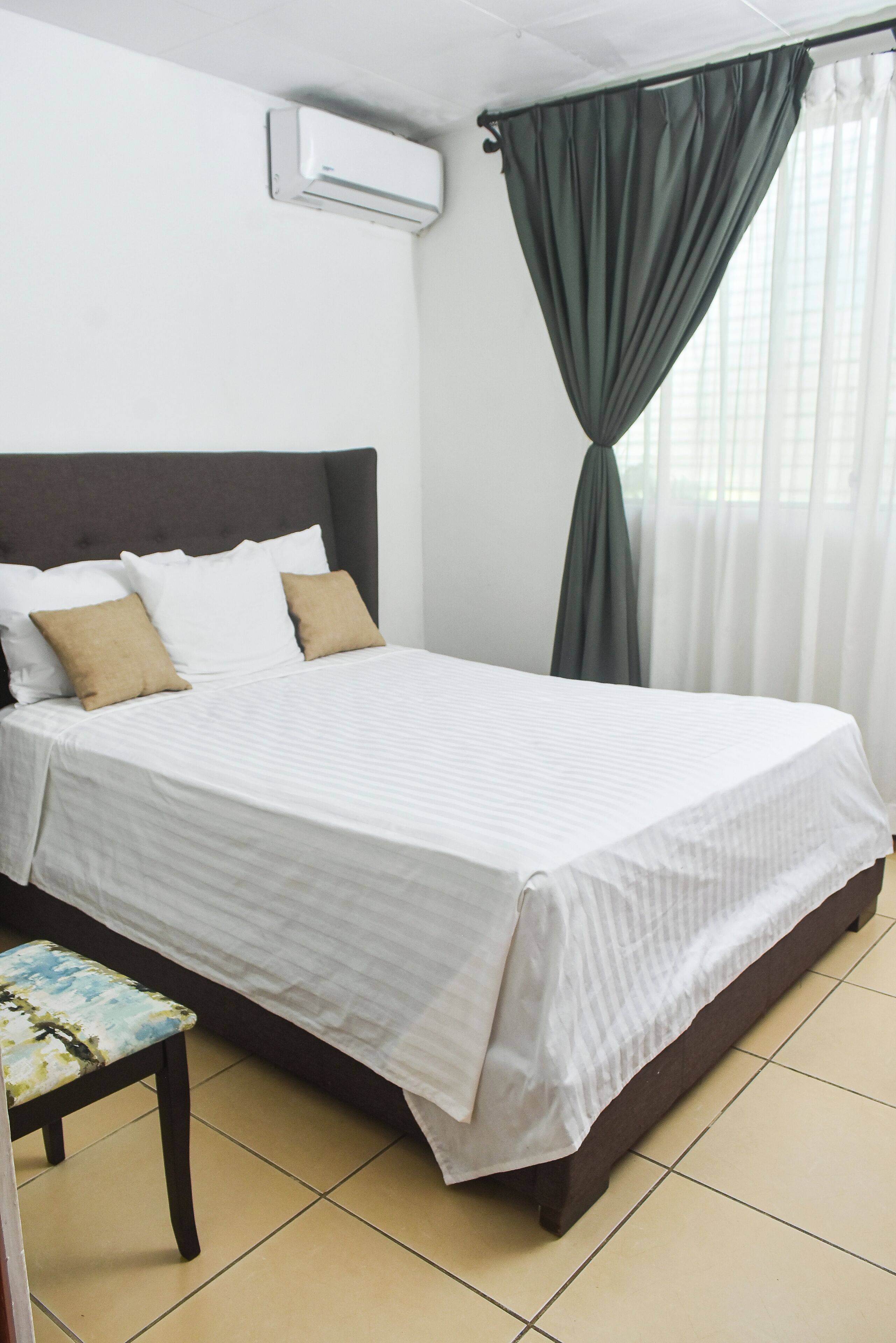 Una cama bien hecha con ropa de cama blanca y dos almohadas, una mesita con un cojín estampado y una ventana con cortinas de encaje.