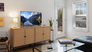Una sala de estar con un televisor de pantalla plana sobre un mueble de madera, una mesa de centro de vidrio, un sofá de cuero negro y una puerta que da a un balcón.