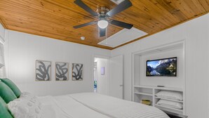 Un dormitorio con cama, ventilador de techo, televisor montado en la pared y cuadros.