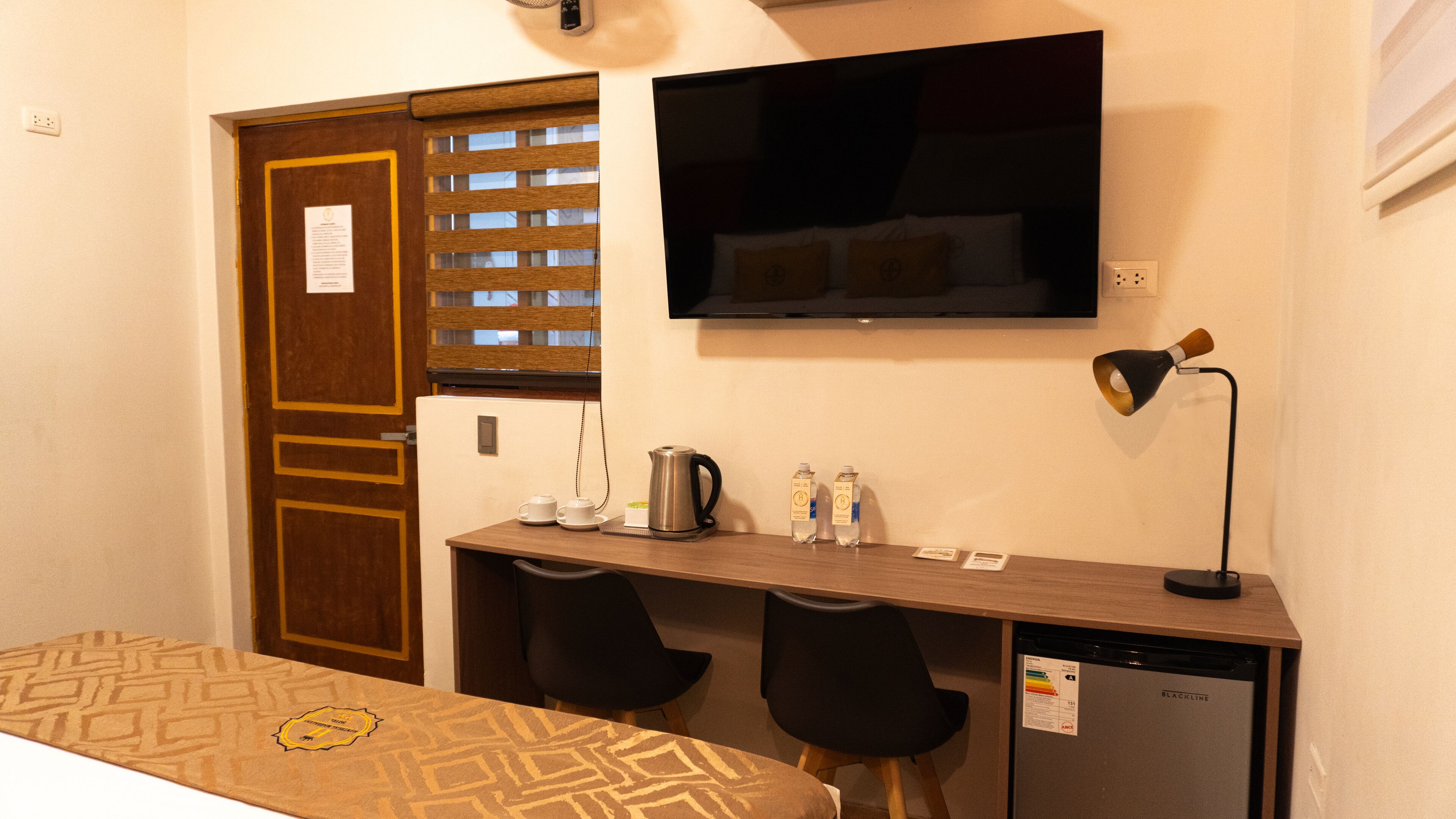 A hotel room with a bed, a desk with two chairs, a coffee maker, a kettle, a TV, a lamp, and a mini fridge.