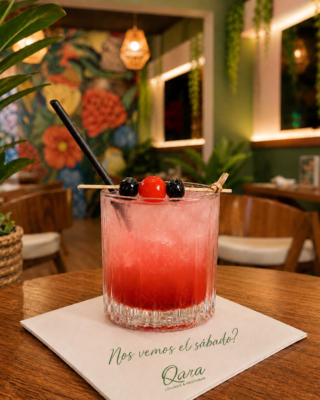 A cocktail with a straw and fruit garnish on a glass, placed on a table with a napkin that reads 'Nos vemos el sábado?' and 'Qara Lounge & Restaurante'.