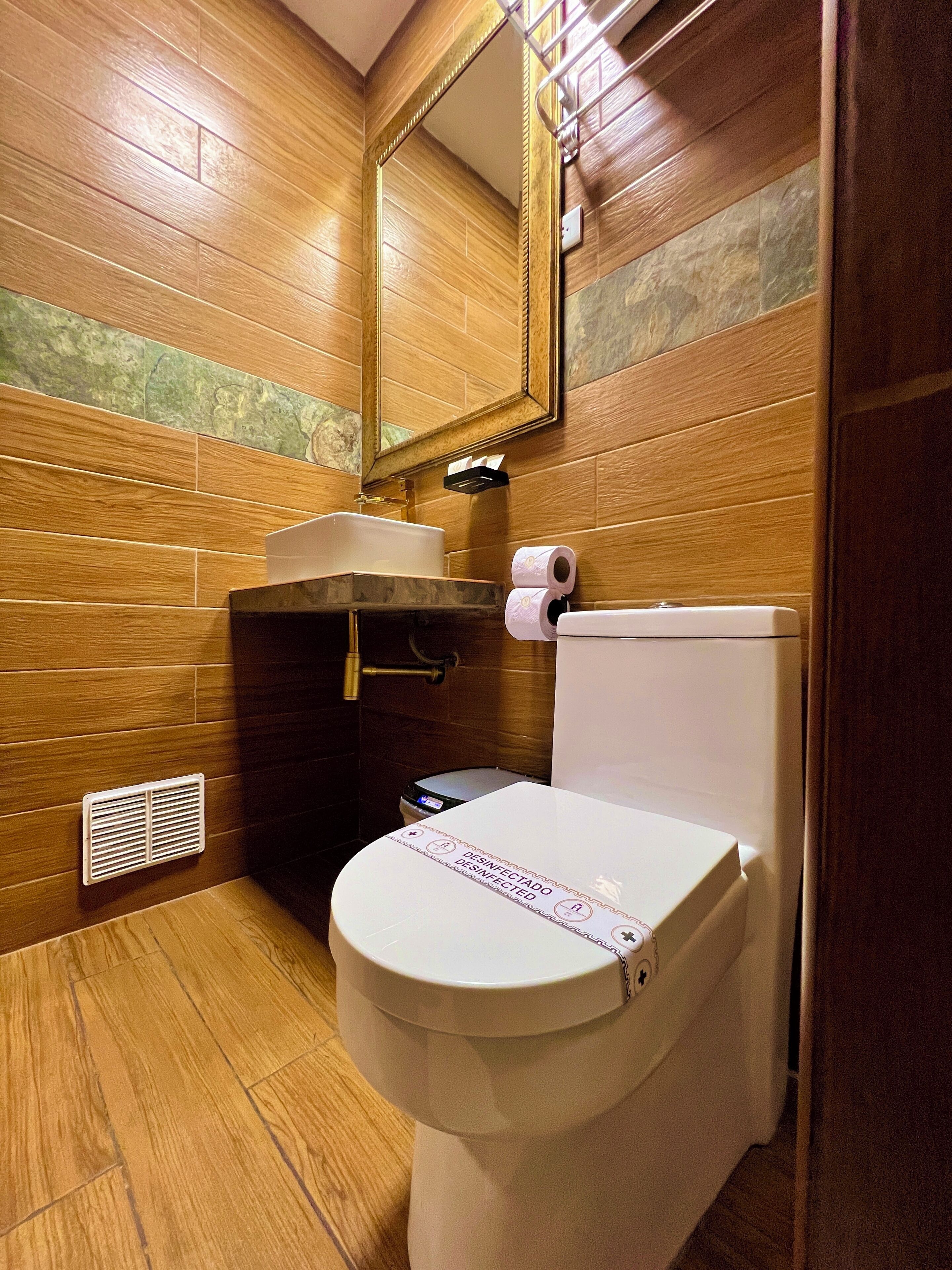 A wooden-finished bathroom with a white toilet, a mirror above the sink, and a vent on the wall.
