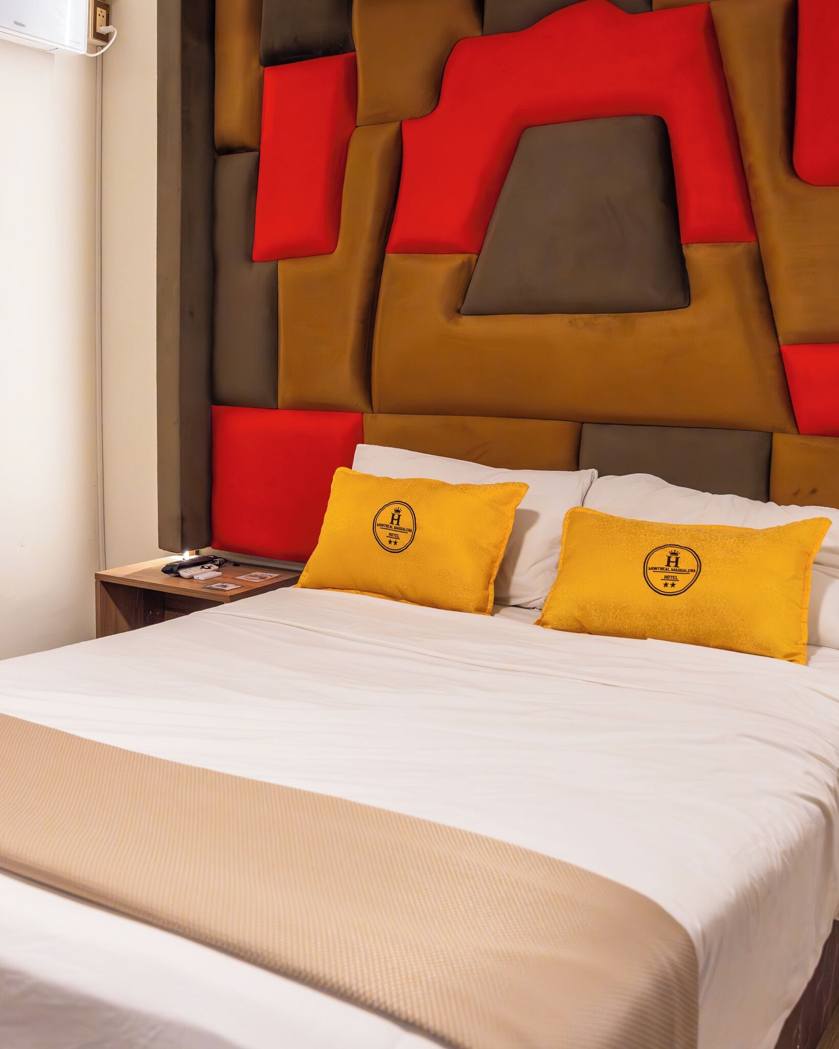 A hotel room with a large bed, two yellow pillows with a logo, a wooden nightstand with a phone and a book, and a modern wall design.
