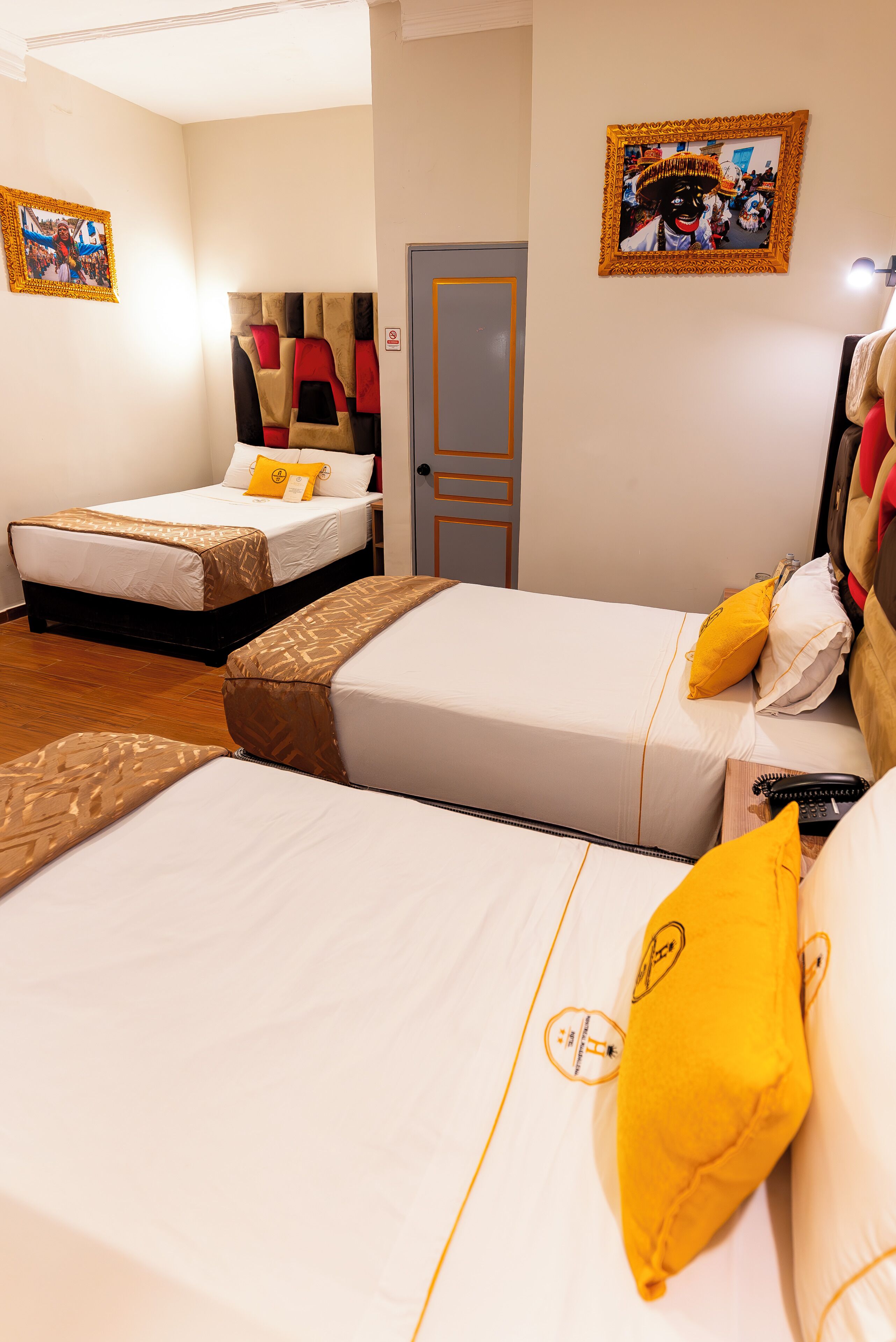 Habitación de hotel con dos camas, cada una con una colcha estampada y almohadas amarillas. Se observa una puerta y un cuadro en la pared.