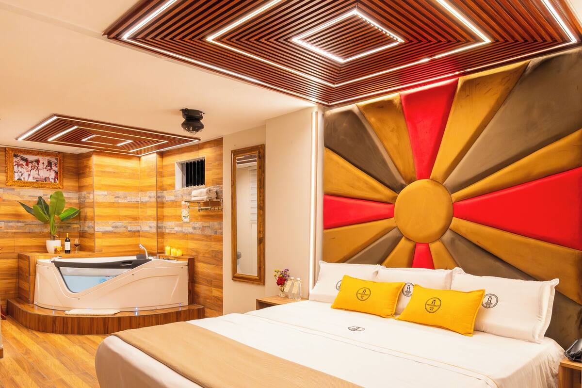 A modern hotel room with a large bed, a built-in bathtub, wooden flooring, and a unique ceiling design.