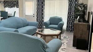 A living room with a sofa, armchair, and coffee table. There is a ceiling fan, a door, and a window with curtains.