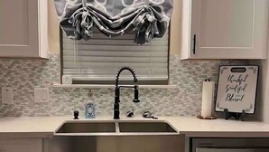 A kitchen with a stainless steel sink, white cabinets, a patterned window valance, and a sign with a positive message.