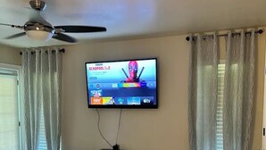 A bedroom with a bed, a ceiling fan, a TV displaying 'DEADPOOL 1 & 2', and a window with curtains.