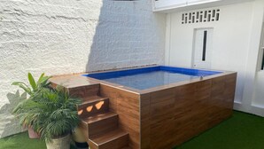Ein rechteckiger Holzpool mit blauem Wasser, umgeben von Kunstgrras und einer weißen, strukturierten Wand.