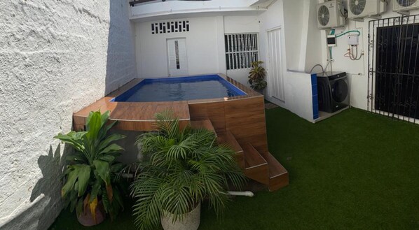 Une piscine dotée d’une terrasse en bois, entourée de gazon synthétique et de plantes en pot.