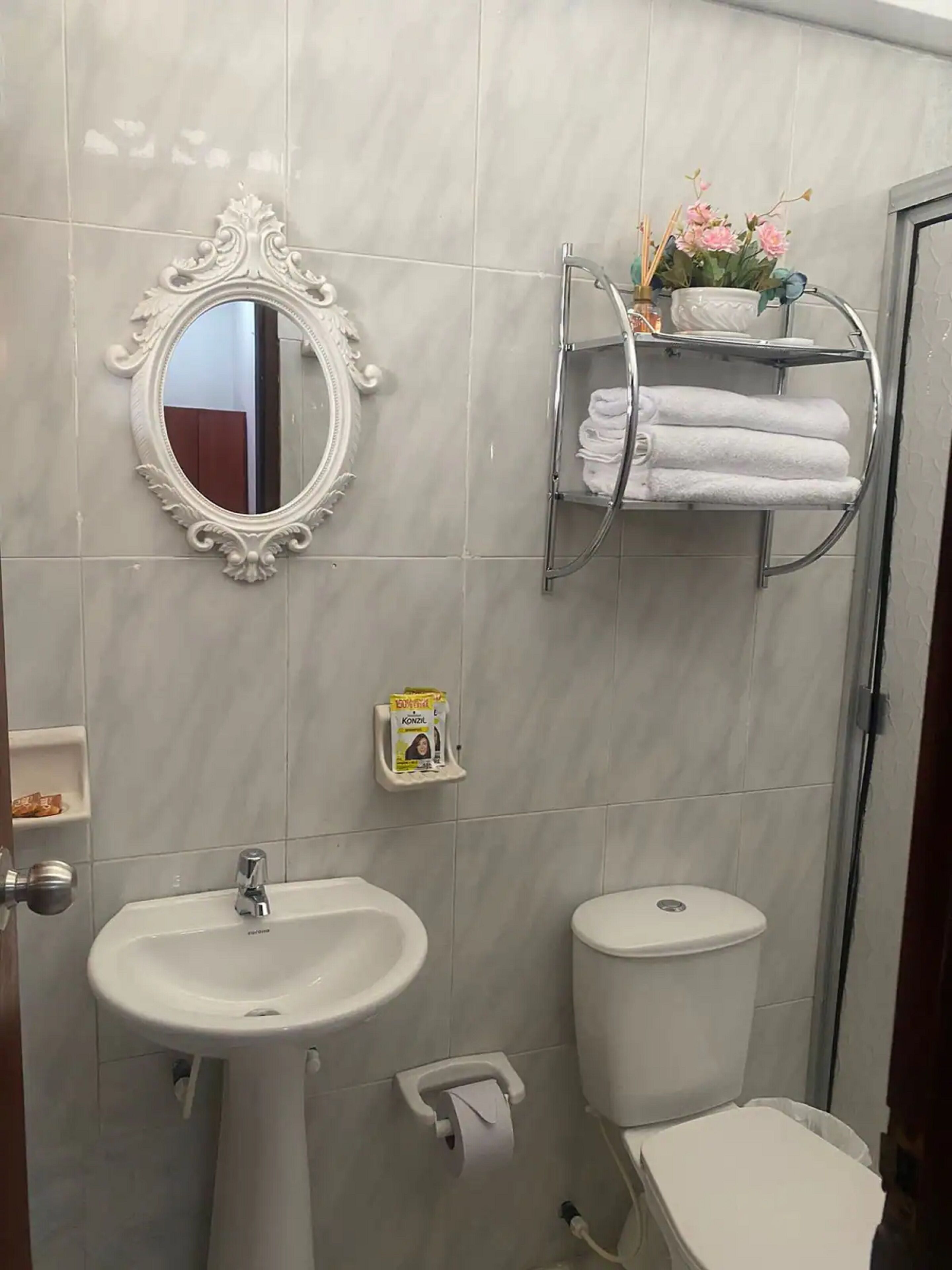 A bathroom with a pedestal sink, toilet, and a mirror with an ornate frame. There is a shelf with towels and a small box of toilet paper.
