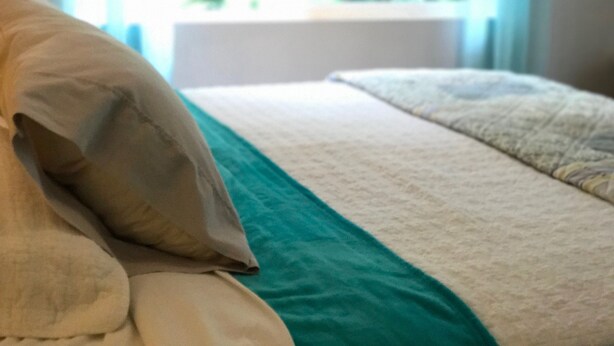 A bed with a teal and white bedspread, a beige pillow, and a window with a view of greenery. - Puolani Iki-Sweet lil cottage near National Park (Volcano)