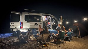 Een groep mensen zit 's avonds aan een tafel op een zandstrand, met een busje met de tekst 'Sampa' op de achtergrond.