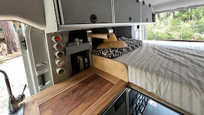 Een compact RV-interieur met een kitchenette, een bed met beddengoed met een patroon, en uitzicht op groen buiten.