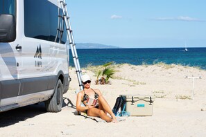 A person sitting on the sand near a vehicle with a ladder, a beach, and the ocean in the background. - Sampa Camper Van Adventure - Explore in Style! (Cabo San Lucas)