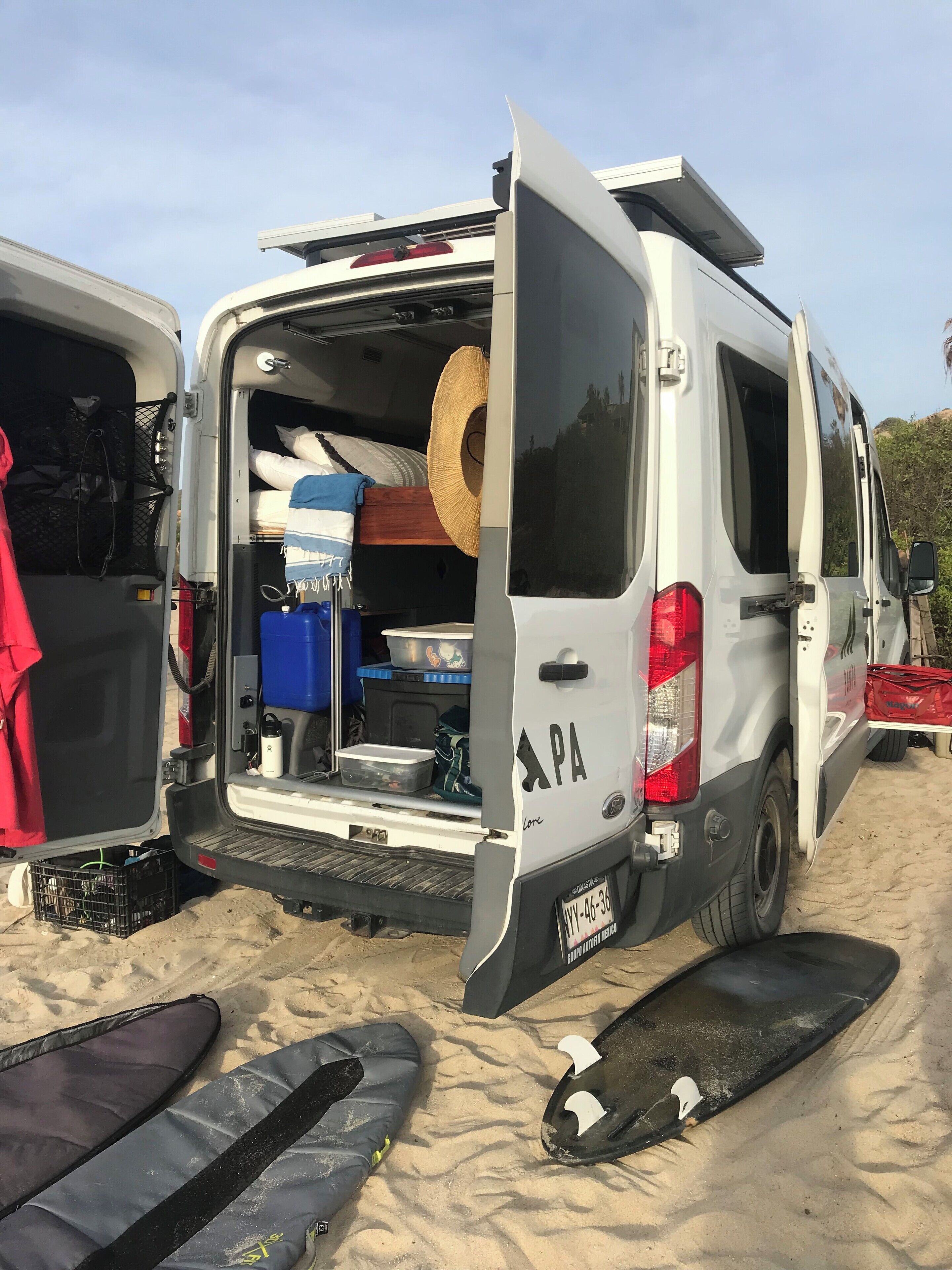 Een witte bestelwagen met open achterdeur, waar een bed, een strohoed, een blauwe koelbox en diverse spullen in liggen.