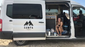 Een witte bus met het logo "SAMPA explore" op de achterkant, geparkeerd op een zandstrand.
