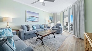 A living room with a sofa, coffee table, and a ceiling fan. There is a large window with curtains and a view of the outdoors. - Palacio 805 | Beachfront | Beachside & Indoor Pool, Hot Tub | My Beach Getaways (Pensacola)