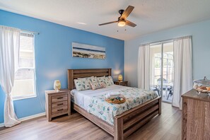 A bedroom with a wooden bed, a ceiling fan, a nightstand with a lamp, and a view of the outdoors through a sliding glass door.