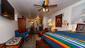 Quarto com uma cama, uma cadeira azul, uma mesa de madeira, um ventilador de teto e uma televisão.