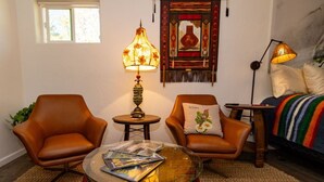 Uma sala de estar aconchegante com duas poltronas de couro, uma mesa de centro de vidro com revistas, um abajur decorativo e uma cama com um cobertor colorido.