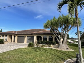 Stucco-clad building with arched windows and a flat roof, set amidst a lush lawn with palm trees and shrubs.