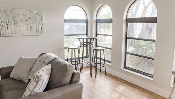 Una habitación con un sofá de cuero marrón, dos ventanas arqueadas, una mesita con taburetes y un cuadro de árboles en la pared.