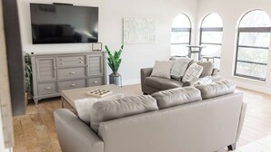 Una sala de estar moderna con un sofá gris, un televisor de pantalla plana, un mueble gris y ventanas grandes.