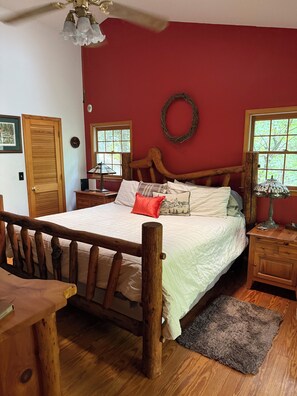 Quarto com cama de madeira, mesa de cabeceira, abajur, janela com vista e porta.