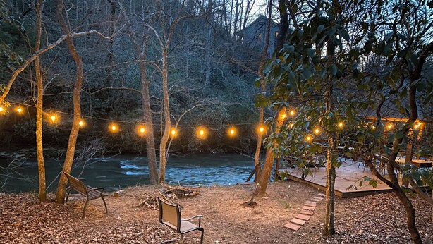 A rustic outdoor seating area with a bench and a wooden deck overlooking a river, surrounded by trees and string lights.
