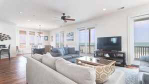 A spacious living room with a large sofa, a coffee table, and a television. There are multiple windows and a balcony with a view of the ocean. - Sweeping Views, Oceanfront Porches, Private Beach Access, Close to Lighthouse - Double Stack (Folly Beach)