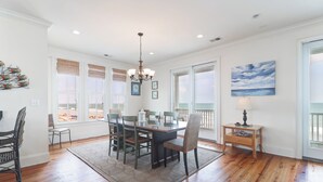 A dining room with a wooden table, chairs, a chandelier, and large windows with blinds. - Sweeping Views, Oceanfront Porches, Private Beach Access, Close to Lighthouse - Double Stack (Folly Beach)