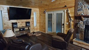 Ein gemütliches Blockhausschlafzimmer mit Holzbalkendecke, einem an der Wand befestigten Flachbildfernseher, einem Kamin aus Naturstein und einer bequemen Sitzecke mit Ledersofa und Sesseln.