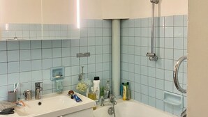 Un baño con lavamanos, espejo y bañera. Hay artículos de aseo en el mostrador y un perchero para toallas.