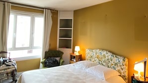 Habitación de hotel con cama, una silla, mesita de noche con lámpara, ventana con cortinas y estante con objetos.