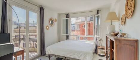 A bedroom with a bed, a desk, a chair, a TV, a wardrobe, and a balcony with a view of buildings.