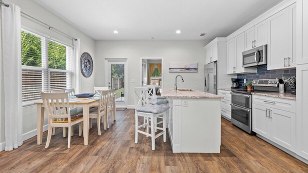 A modern kitchen with white cabinetry, a central island, and a dining area with wooden chairs. - New Home 3/2 With Garage Near Downtown (Mount Dora)